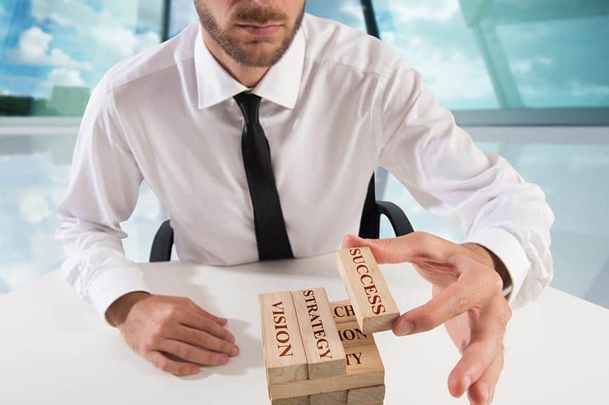 A businessman building a Jenga stack with inscribed blocks: Vision, strategy, success