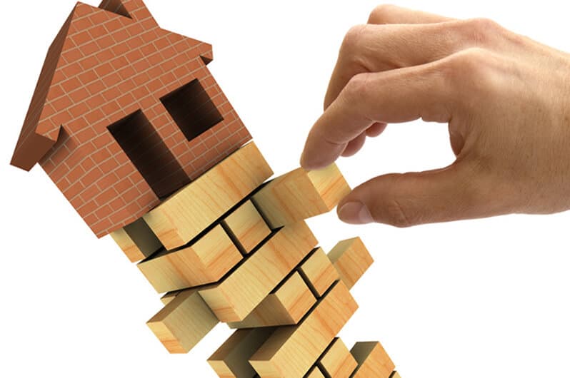 A hand removing blocks from a Jenga stack with a house on top of it.